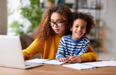 Genç ve mutlu oğlu olan pozitif bir anne masada oturup dizüstü bilgisayar kullanıyor, ekrana bakıyor ve birlikte komik videolar izlerken gülümsüyor. Afro-amerikan ailesi evde vakit geçiriyor.