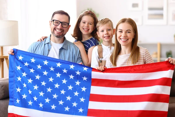 若い幸せなアメリカの家族の両親と2人の小さな子供たちは 独立記念日を祝いながら 米国の国旗とカメラで笑顔で自宅でソファに座っています 愛国的なアメリカの休日の概念 — ストック写真