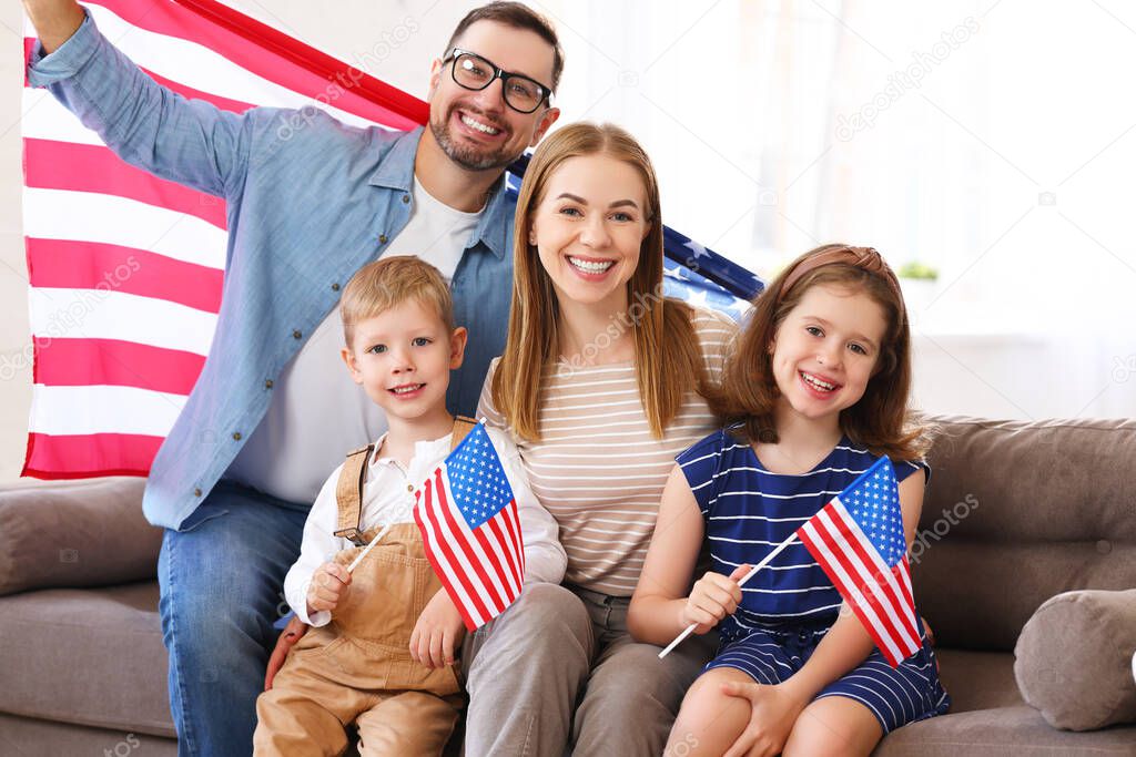 Young happy american family parents and two little kids sitting on sofa at home with flags of united states and smiling at camera while celebrating Independence Day. Patriotic US holiday concept