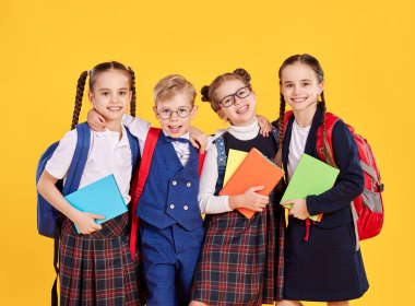 Okul üniformalı, sırt çantalı, sarı arka planda yan yana kameraya bakan iyimser sınıf arkadaşları.