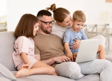 Genç ve mutlu bir aile evde dinleniyor. Baba, anne ve iki çocuk kanepede otururken dizüstü bilgisayar kullanıyor, çocukları olan ebeveynler, oğulları ve kızları birlikte video izliyor ya da internette geziniyorlar.