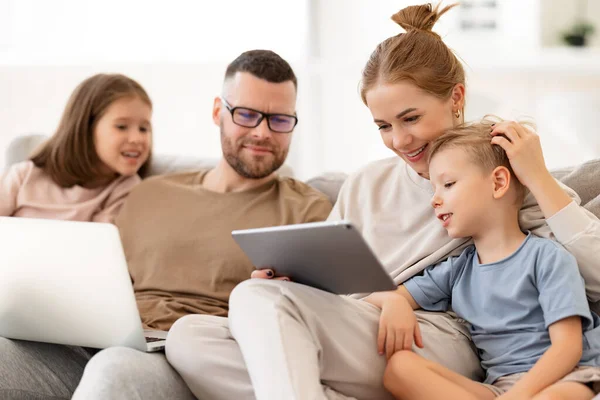 年轻快乐的家庭 有两个孩子的父母在使用现代技术的同时一起呆在家里 带着笔记本电脑 数码平板电脑和智能手机的父亲 母亲和孩子们在沙发上放松一下 — 图库照片