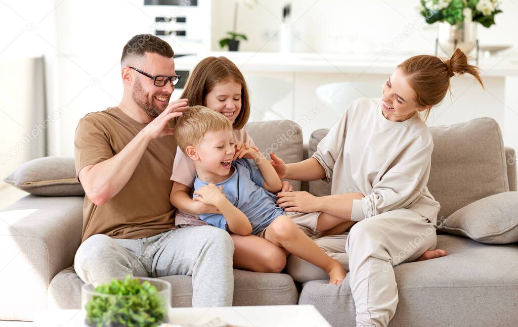 Happy family, young parents with two cute excited children son and daughter having fun, embracing and playing together while relaxing on sofa at home. Mom, dad and kids enjoying leisure time