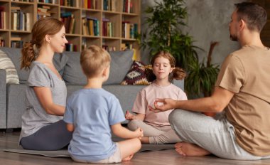 Genç ebeveynler evde çocuklarla meditasyon yapıyor. Güzel aile babası, anne ve gözleri kapalı iki çocuk. Lotus poz verirken yoga yapıyorlar.