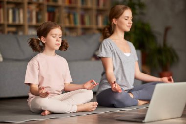 Bilgisayarla yoga yapan bir aile. Genç kadın anne ve küçük kız günlük giysiler giyip birlikte meditasyon yaparken oturma odasındaki dizüstü bilgisayarın önünde poz veriyorlar.