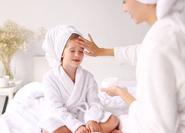 在家里的日常护肤活动中 穿着浴衣 双腿交叉坐在床上的女人在女孩的额头上涂奶油 — 图库照片