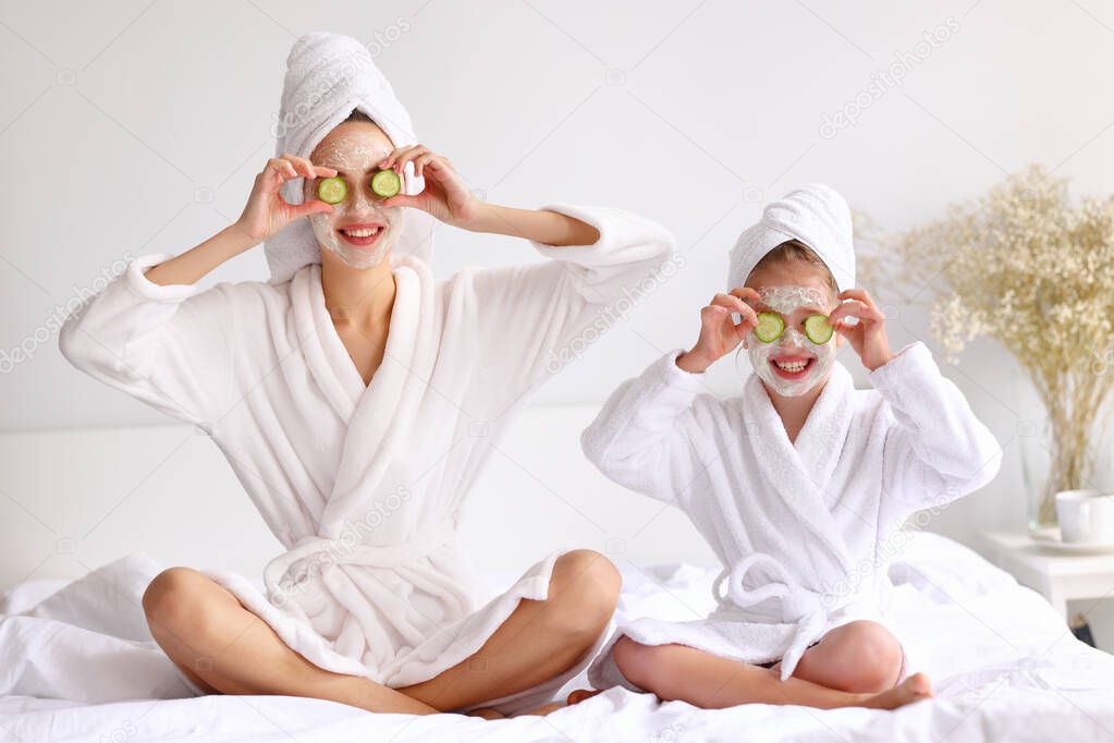 Optimistic woman and girl with moisturizing mask smiling and holding cucumber slices near eyes while sitting cross legged on bed during skin care procedure