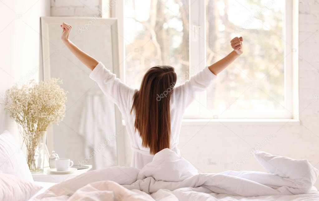 Back view of anonymous woman in sleepwear raising arms and stretching body while sitting on bed in morning in light bedroom