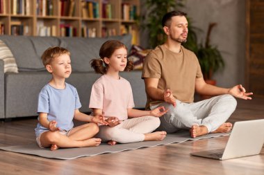 Bilgisayarla yoga yapan bir aile. Genç adam, baba ve küçük çocuklar birlikte meditasyon yaparken Lotus 'ta oturmuş oturma odasında dizüstü bilgisayarın önünde poz veriyorlar.