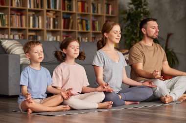 Genç ebeveynler evde çocuklarla meditasyon yapıyor. Güzel aile babası, anne ve gözleri kapalı iki çocuk lotus poz verirken yoga yapıyorlar.