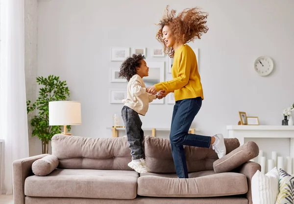 Crazy weekend with mom. Joyful afro american family mother and little son holding hands and jumping on sofa in living room at home, full length shot. Mom and child in casual wear playing together