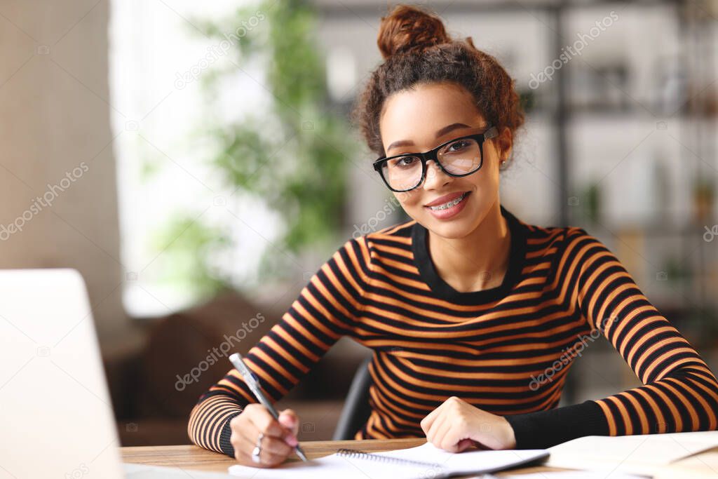 Beautiful young afro american business woman analyzing documents while sitting at her workplace and working remotely on laptop. Happy mixed race female student studying online during covid 19 outbreak