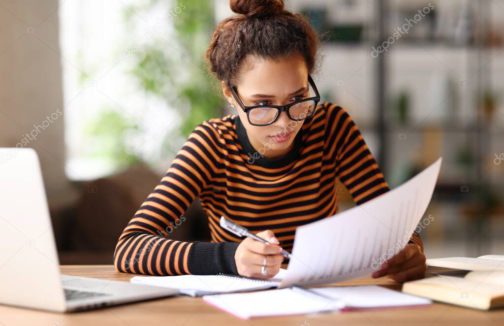 Beautiful young afro american business woman analyzing documents while sitting at her workplace and working remotely on laptop. Happy mixed race female student studying online during covid 19 outbreak