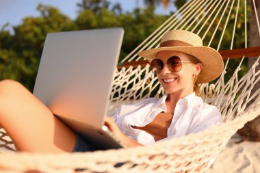 Uzaktan çalışıyor. Genç mutlu kadın, hasır şapkalı ve güneş gözlüklü kadın güneş gözlüklü, gün batımında plajda hamakta dinlenirken. Tatil sırasında uzaklık işi
