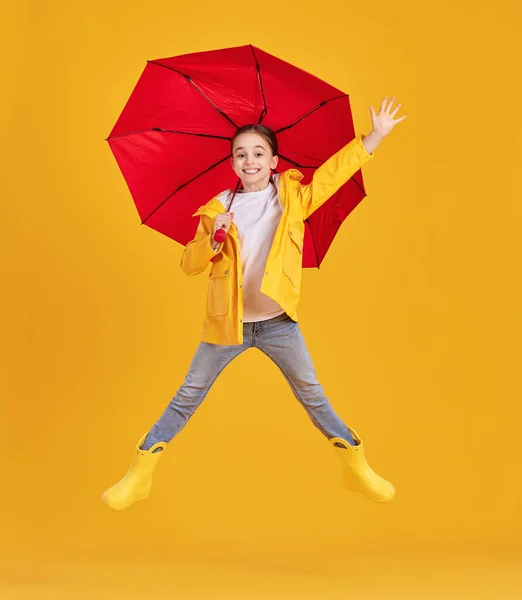 フルボディサイドビューの興奮した女の子で黄色のレインコートとブーツとともに赤い傘持っています楽しいですとジャンプ上地面に対して黄色の背景 — ストック写真