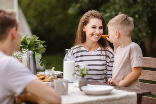 Family breakfast in nature in the garden. Cheerful mom and little son   with dad drink milk and eat fresh cookies on a summer morning at the table