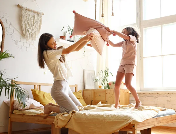 Happy family morning. Full length of overjoyed excited young mother and excited little daughter having pillow battle on bed, two girls older and younger sister laughing having fun in bedroom at home