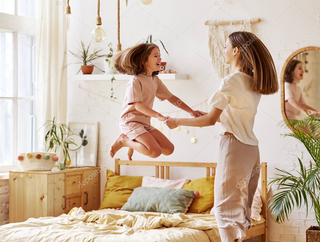 Feeling carefree. Excited cute little preschool girl spending time with young mother at home, happy child jumping on bed and looking at camera. Younger and older caucasian sister having fun together