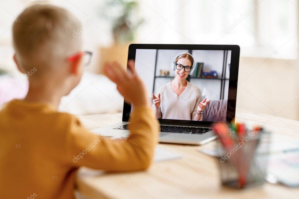 Happy child boy in headphones smiling and waves his hand to the teacher as a sign of greeting while making video call   during online lesson at home