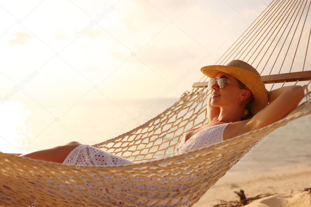 Enjoying summer vacation. Young happy woman wearing straw hat and sunglasses keeping eyes closed and relaxing while lying in the hammock on the sandy beach at sunset. People traveling concept