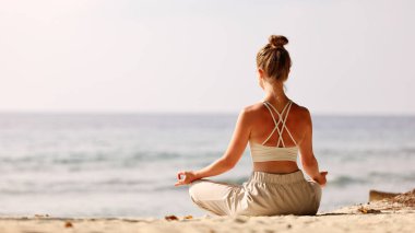 Padmasana 'da gün batımında deniz kıyısında gökyüzüne karşı meditasyon yapan bir kadının arka görüntüsü.