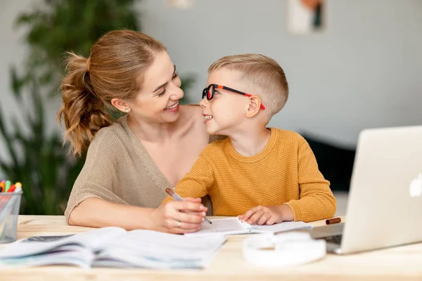 快乐的小男孩 戴着耳机 一边在抄本里做练习 一边在家里通过笔记本电脑远程学习 快乐的妈妈坐在旁边给予支持 — 图库照片