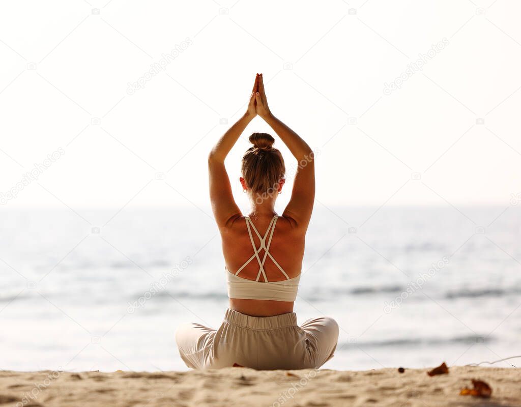 Back view of female sitting in Padmasana while meditating alone on seashore against sky in sundown light
