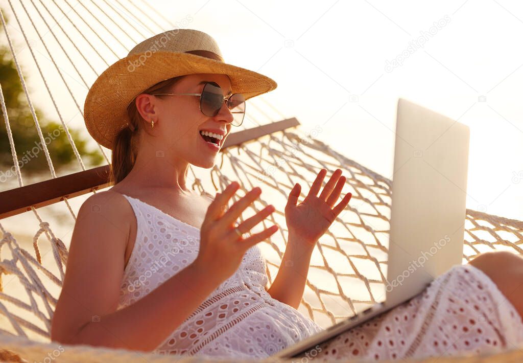 Having online conversation. Young happy successful woman wearing straw hat gesturing   and smiling while having video call on laptop, relaxing in the hammock on tropical sandy beach on sunny day