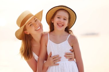 Mutlu bir kadın neşeli bir kıza sarılıyor ve kameraya bakıyor. Bulanık arka planda gün batımı gökyüzü ve deniz tatil köyünde.