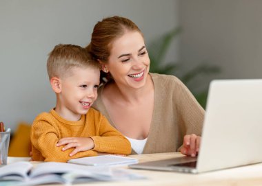 Kulaklık takan neşeli küçük okul çocuğu evde dizüstü bilgisayarla eğitim görürken yanında mutlu annesi otururken ve destek verirken...