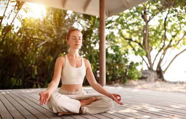 Lotus 'ta çıplak ayaklı genç bir kadın terasın ahşap zemininde poz veriyor ve yazın plajda yoga sırasında gözleri kapalı meditasyon yapıyor.