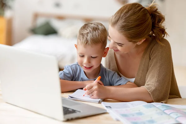 快乐的小男孩 戴着耳机 一边在抄本里做练习 一边在家里通过笔记本电脑远程学习 快乐的妈妈坐在旁边给予支持 — 图库照片