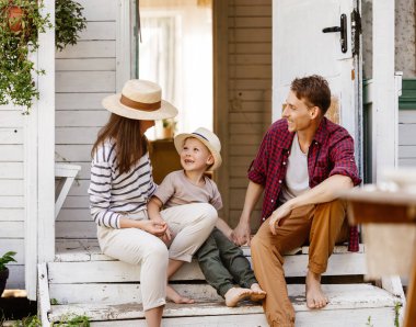 Bütün vücut çıplak ayakla, anne-baba ve oğul gülerken verandada, merdivenlerde oturuyor ve kırsal kesimde yaz günü evin dışında dinleniyor.