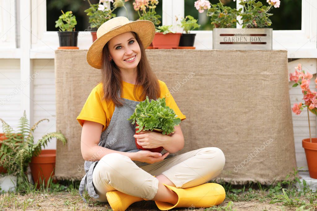 Delighted female gardener with potted plant smiling and looking at camera while sitting cross legged outside orangery