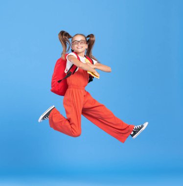 Mutlu, enerjik, günlük kıyafetler giyen, sırt çantası ve el kitabı tutan ve mavi arka planda yükseğe zıplayan bir ilkokul öğrencisiyle dolu.