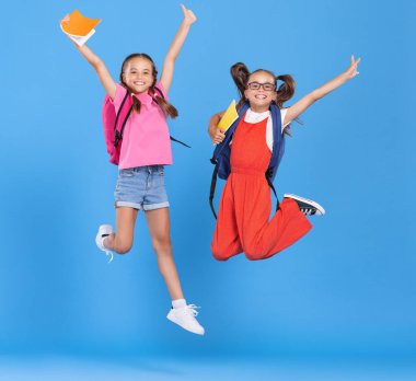 Sırt çantası ve el kitabı tutan ve mavi arka planda yükseğe zıplayan sıradan kıyafetler giyen iki mutlu ve enerjik ilkokul çocuğunun tüm vücudu.