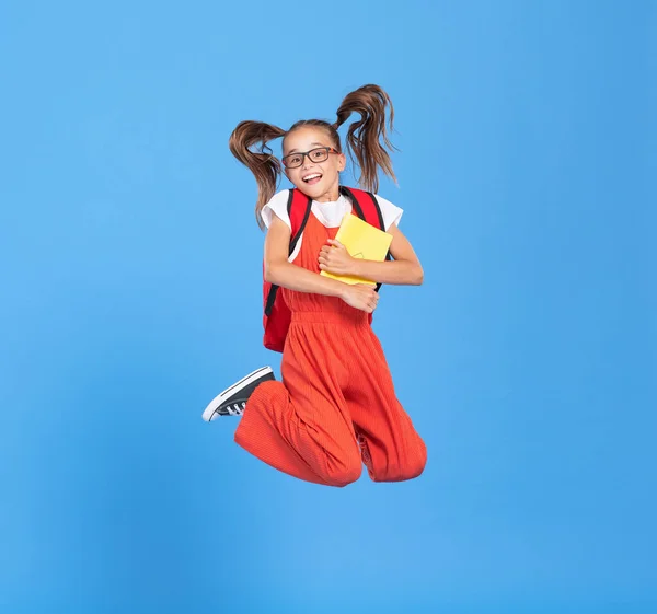 Full body of happy energetic primary schoolgirl  wearing casual clothes holding backpackand copybook in hands and jumping high against blue background