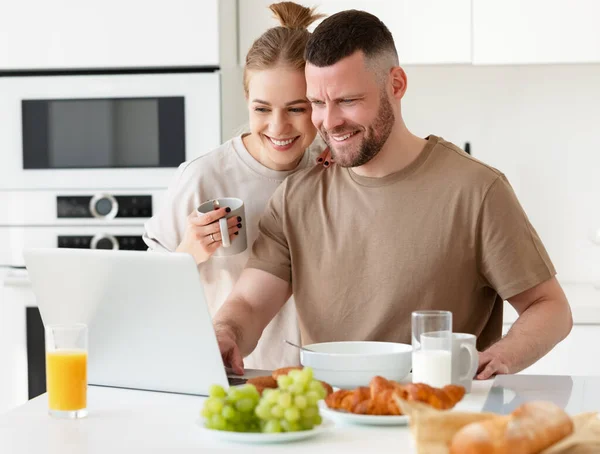 朝一緒に朝食を楽しみながらノートパソコンの画面を見ている若い美しい幸せな家族のカップルは 白いモダンなキッチンに立っています 妻と夫は食事をしながらオンラインでニュースを読む — ストック写真