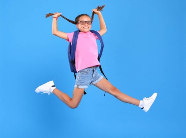 Okula dönmek için acele et. Mavi stüdyo arka planında mutluluk ve sevinçten atlayan, gözlüklü, komik, şaşırmış yüz ifadesiyle enerjik bir okul kızının tam boy fotoğrafı.