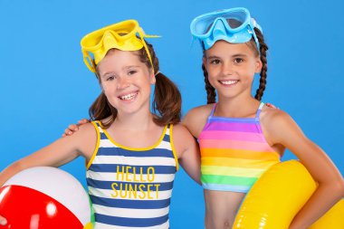 İki küçük kız mayo giymiş en iyi arkadaş lastik halkalı, rüzgar topu ve renkli yüzme gözlükleriyle poz veriyor, beline sarılıyor ve geniş bir gülümsemeyle deniz macerasına hazırlanıyor.