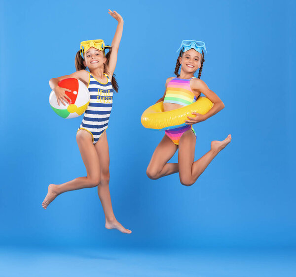 Две забавные маленькие девочки-сестры в купальниках и плавательных очках, прыгающие в воздухе с надувным плавательным кольцом и мячом в руках на синем студийном фоне, взволнованные и готовые к летнему морскому отдыху