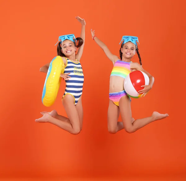 两个穿着泳衣 戴着泳镜 手拿着充气泳圈 手拿着球 手拿着橙色工作室背景的小女孩兴奋地跳起来 准备放暑假 — 图库照片
