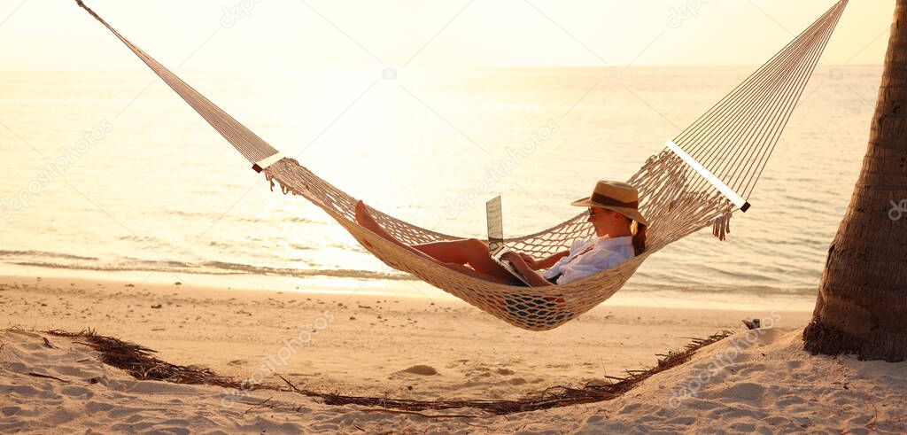 Young woman, successful female freelancer using laptop while lying in hammock on the tropical beach at sunset, working remotely during summer vacation. Distance work concept
