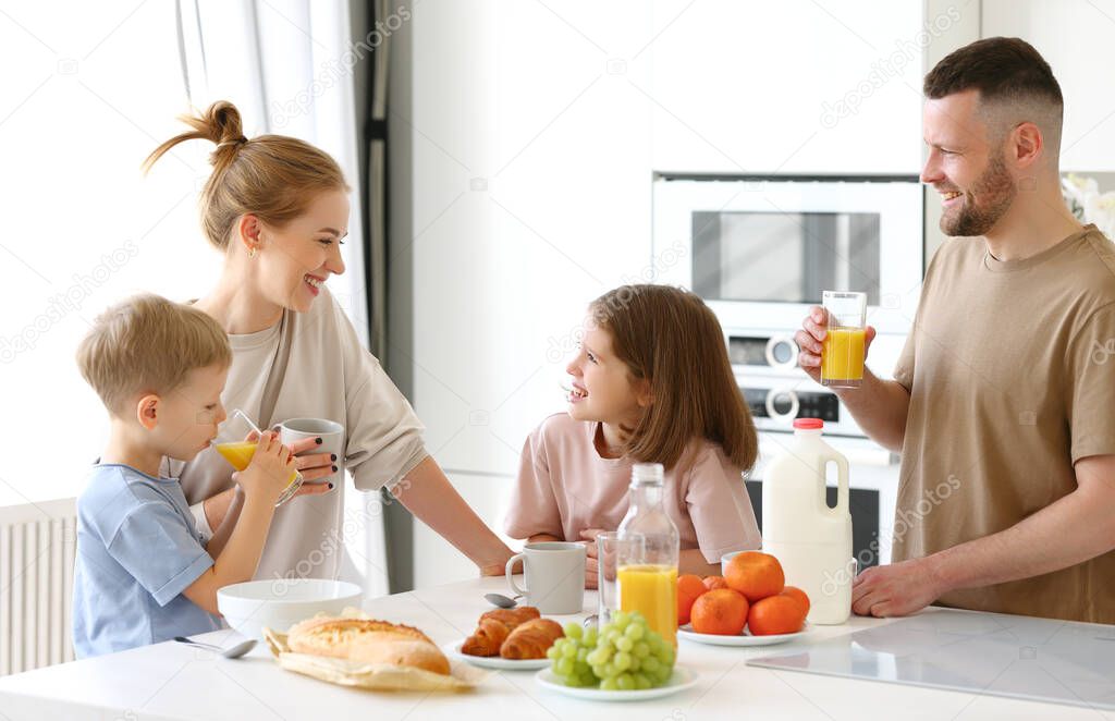 Young happy beautiful family having breakfast together at home. Father, mother and two cute little kids eating healthy food in morning, talking and smiling while standing in modern kitchen