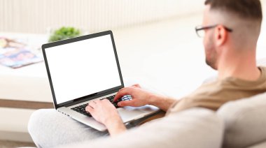 Dizüstü bilgisayar klavyesinde yazan erkek serbest yazar, kanepede oturuyor ve uzaktan çalışıyor, genç adam boş beyaz ekranla çalışıyor. Ekranda seçmeli odak