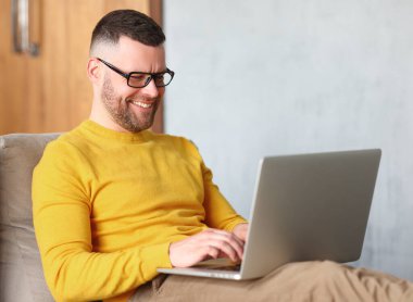 Mutlu genç adam gözlük takıyor rahat bir pozisyonda oturuyor evdeki koltukta oturuyor ve bilgisayar başında çalışıyor, internette sörf yapıyor, sohbet ediyor ya da evde dinlenirken internetten alışveriş yapıyor.