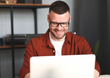 Gözlüklü başarılı bir erkek, uzaktan kumandalı dizüstü bilgisayar kullanıyor, rahat iş yerinde uzaktan çalışmaktan zevk alıyor, klavyeyle yazı yazıyor ve gülümsüyor. Serbest konsept