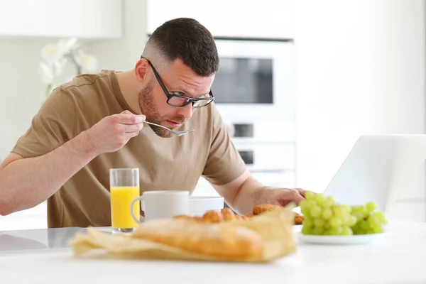 若いです焦点を当てたハンサムな男は 自宅でモダンなキッチンのテーブルに座っている間 ノートパソコン上でリモートで作業眼鏡を身に着けています 男性フリーランス健康的な食事の朝食とコンピュータを見て — ストック写真