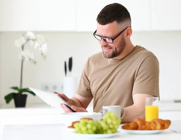家でモダンなキッチンのテーブルに座っている間 若い笑顔ハンサムな男は タブレットの上でリモートで作業眼鏡をかけて 男性のフリーランス健康的な食事の朝食とコンピュータを見て — ストック写真