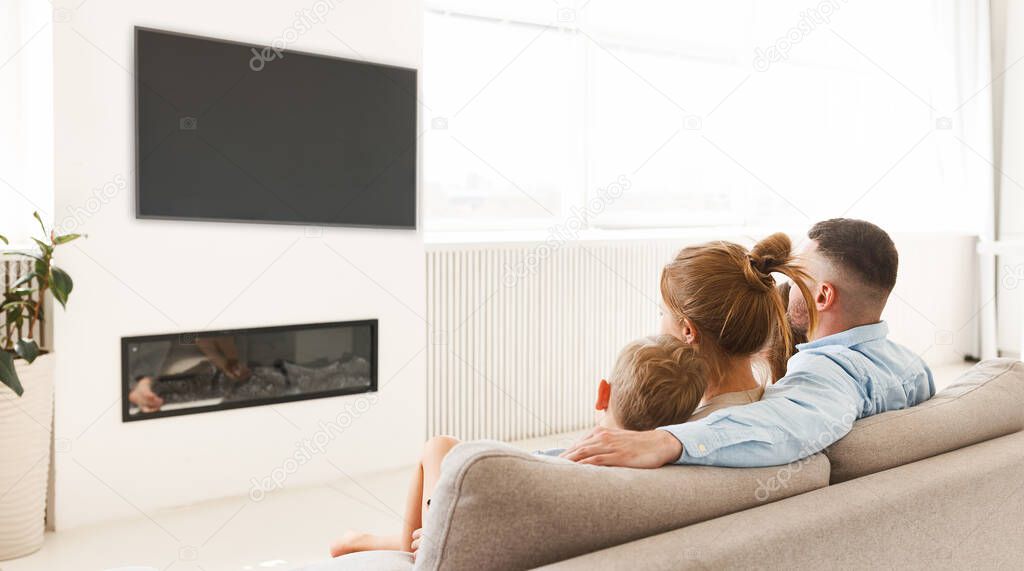 Rear view of young family, father, mother and two little kids sitting on sofa in front of fireplace and watching tv, bonding and enjoying leisure time together at home